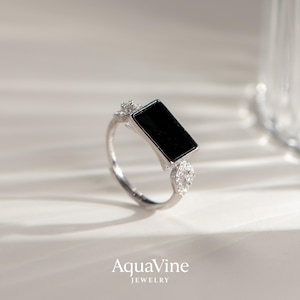 May include: A silver ring with a rectangular black stone at the center, flanked by smaller, clear stones. The ring band is thin and polished. The brand name "AquaVine JEWELRY" is displayed at the bottom.