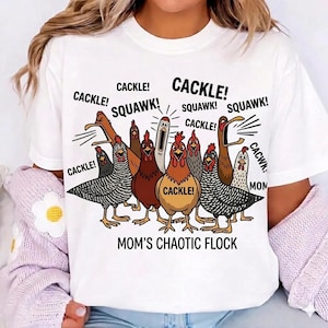 May include: White t-shirt with a cartoon illustration of a flock of chickens. The text includes "CACKLE! SQUAWK!" and "MOM'S CHAOTIC FLOCK". The chickens are in different colours and poses, with speech bubbles.