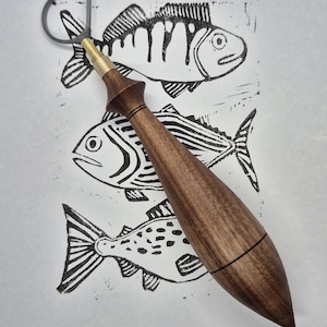 Puede incluir: Una herramienta de cerámica de madera con un lazo de metal se coloca sobre un papel blanco con tres impresiones de peces negros. La herramienta tiene un mango marrón oscuro y un accesorio de latón. Las impresiones de peces varían.