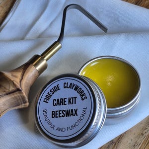 Puede incluir: Un kit de cuidado Fireside Clayworks con cera de abejas en una pequeña lata abierta. Una herramienta de metal y madera está al lado de la lata. El texto en la lata dice "Beautiful and Functional". La cera de abejas es amarilla.