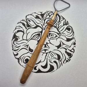 Peut inclure: Un outil de sculpture en argile à manche en bois avec une boucle en métal est positionné sur une impression linogravée en noir et blanc d'un visage de lion stylisé. L'impression est circulaire, et l'outil est incliné dessus.