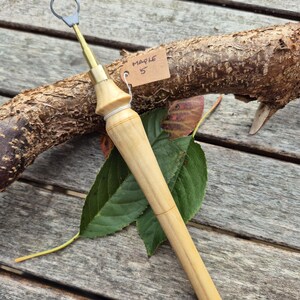 Può includere: Un utensile da ceramica in legno chiaro con un anello di metallo all'estremità. L'impugnatura è in legno chiaro e una piccola etichetta recita "MAPLE 5". L'utensile è appoggiato su una superficie di legno con foglie e un ramo.