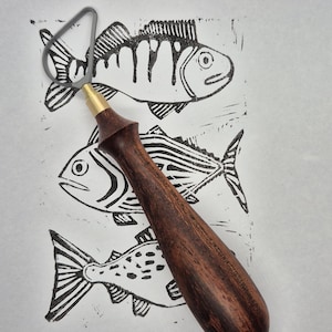 May include: A linocut print of three fish in black ink on white paper, with a dark wooden handled linocut carving tool. The tool has a brass ferrule and a gray metal blade. The fish are stylized with detailed lines.