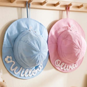 May include: Two sun hats, one light blue and one light pink, each featuring a wide brim and a rounded crown. The blue hat displays the name Willow in white cursive lettering with a small flower, while the pink hat displays the name Olivia.