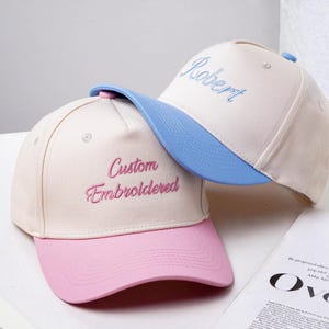 Könnte beinhalten: Zwei Baseballkappen mit individueller Stickerei. Eine Kappe ist cremefarben mit rosa Schirm und dem Schriftzug "Custom Embroidered" in Rosa. Die andere ist cremefarben mit blauem Schirm und dem Namen "Robert" in Blau gestickt.