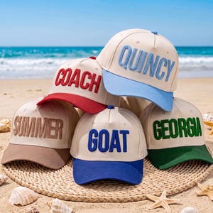 Puede incluir: Una colección de cinco gorras de béisbol de color crema con coronas estructuradas y viseras en contraste. Cada gorra presenta letras grandes, bordadas y en relieve en diferentes colores: COACH en rojo, QUINCY en azul claro, SUMMER en beige, GOAT en azul y GEORGIA en verde.