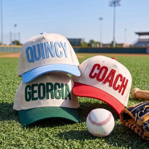 Könnte beinhalten: Drei Baseballkappen mit cremefarbenen Kronen und farbigen Schirmen. Eine Kappe zeigt den hellblauen Schriftzug "QUINCY" mit hellblauem Schirm, eine den grünen Schriftzug "GEORGIA" mit grünem Schirm und eine den roten Schriftzug "COACH" mit rotem Schirm.