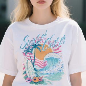 May include: White t-shirt with a colorful graphic design. The design features a sunset, palm trees, a surfboard, and a wave. The text "Sunset Chaser" is written across the top in a script font.