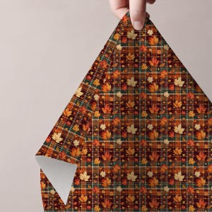 May include: A sheet of wrapping paper featuring a dark brown, orange, and green plaid pattern overlaid with a repeating print of autumn maple leaves in shades of yellow, orange, and red. The paper is folded at the top corner to reveal a white underside.