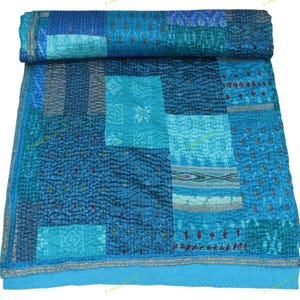 Op de afbeelding: Een patchworkquilt in tinten blauw, blauwgroen en turquoise. De quilt heeft een verscheidenheid aan patronen van vierkanten en rechthoeken, aan elkaar gestikt met zichtbare draad. De randen zijn afgewerkt met een effen blauwe rand.