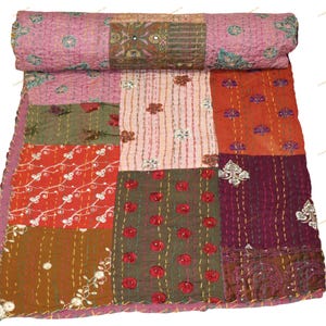 May include: A patchwork quilt with a rolled top, featuring a variety of colors and patterns. The quilt is made up of squares in shades of pink, red, orange, green, and brown, with embroidered floral and mirror accents. The quilt is a decorative textile.