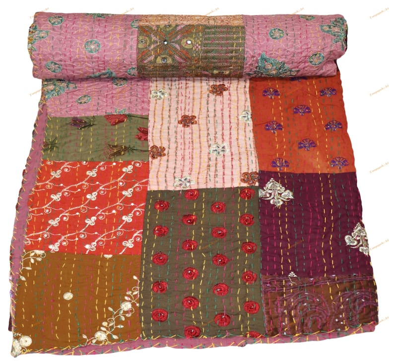 May include: A patchwork quilt with a rolled top, featuring a variety of colors and patterns. The quilt is made up of squares in shades of pink, red, orange, green, and brown, with embroidered floral and mirror accents. The quilt is a decorative textile.
