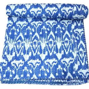 Op de afbeelding: Een opgerolde blauw-witte quilt met patroon. De quilt heeft een herhalend ikat-ontwerp in blauw en wit. De randen zijn netjes gestikt met witte draad en de quilt is opgevouwen.