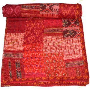 Könnte beinhalten: Ein Patchwork-Quilt in Rottönen mit einem aufgerollten Abschnitt. Der Quilt zeigt verschiedene Muster und Texturen, darunter geometrische Formen und florale Designs. Die Gesamtästhetik ist warm und einladend, geeignet für ein Bett oder Sofa.