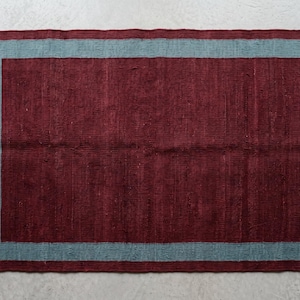 Peut inclure: Un tapis rectangulaire présentant un champ central bordeaux profond entouré d'une large bordure bleu sarcelle terne. Le tapis a un aspect tissé et texturé, fini avec des franges courtes de couleur claire sur les deux bords verticaux.