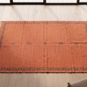 May include: A rectangular area rug in a burnt orange hue featuring a dark charcoal border with a repeating triangular pattern. The rug includes small, centered geometric symbols in dark charcoal and is finished with white fringe on two ends.