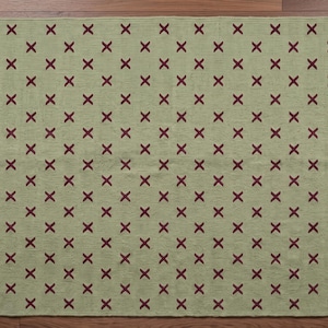 Puede incluir: Una alfombra rectangular con un fondo verde salvia claro y un patrón repetitivo de pequeñas marcas en forma de X de color granate oscuro dispuestas en cuadrícula. La alfombra tiene una textura de tejido plano y bordes acabados.