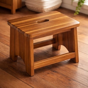 May include: A small, rectangular wooden step stool with a natural finish. The stool has a handle cut-out on the top and a lower support bar. The wood grain is visible, and the stool is designed for use in a variety of settings.