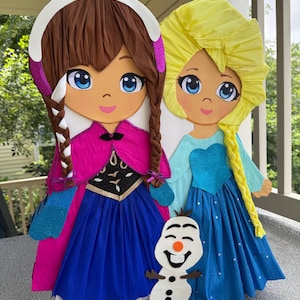 Puede incluir: Dos grandes y coloridas mu&ntilde;ecas de papel de personajes de dibujos animados, una con trenzas marrones y un vestido rosa y azul, la otra con pelo rubio y un vestido azul. Una peque&ntilde;a figura de mu&ntilde;eco de nieve est&aacute; delante de ellas.