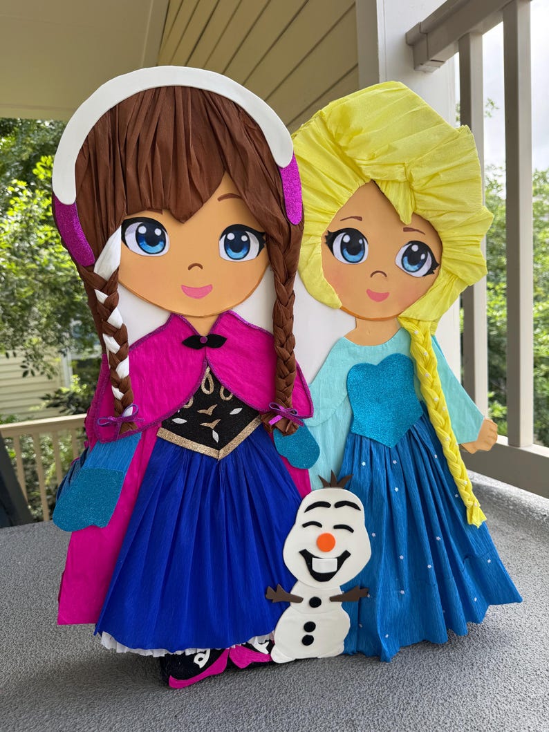 Puede incluir: Dos grandes y coloridas mu&ntilde;ecas de papel de personajes de dibujos animados, una con trenzas marrones y un vestido rosa y azul, la otra con pelo rubio y un vestido azul. Una peque&ntilde;a figura de mu&ntilde;eco de nieve est&aacute; delante de ellas.