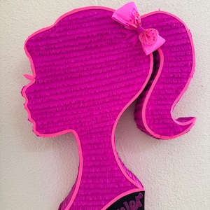 May include: A magenta piñata in the shape of a woman's silhouette, with a pink bow and the name "River" on the base. The piñata is made of fringed paper and has a bright pink outline.