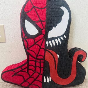 May include: A handmade pi&ntilde;ata featuring a split design of Spider-Man and Venom. The Spider-Man side is red with black webbing and a white eye. The Venom side is black with a white mouth and teeth, and a red glitter tongue.
