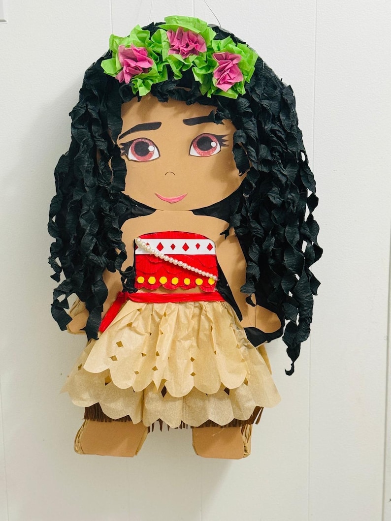 May include: A handmade pi&ntilde;ata in the shape of a cartoon character. The character has black paper hair, a red and white top, and a tan skirt. A green and pink floral crown sits atop the head. The pi&ntilde;ata is designed to resemble a popular animated character.