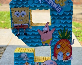 May include: A number nine piñata featuring characters from the animated series SpongeBob SquarePants. The piñata is blue and yellow with images of SpongeBob, Patrick Star, Squidward, Gary, and the pineapple house. The text "Bob Esponja" is also visible.