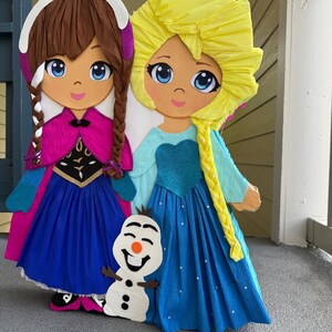 Puede incluir: Dos grandes y coloridas figuras de papel mach&eacute; de personajes animados, una con trenzas marrones y capa rosa, la otra con trenza amarilla y vestido azul. Una peque&ntilde;a figura de mu&ntilde;eco de nieve se encuentra entre ellas. Adecuado para una fiesta infantil.