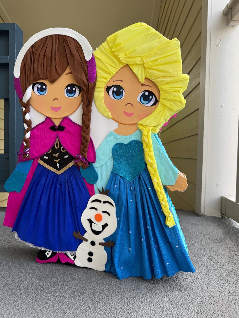 Puede incluir: Dos grandes y coloridas figuras de papel mach&eacute; de personajes animados, una con trenzas marrones y capa rosa, la otra con trenza amarilla y vestido azul. Una peque&ntilde;a figura de mu&ntilde;eco de nieve se encuentra entre ellas. Adecuado para una fiesta infantil.
