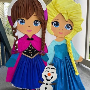 Puede incluir: Dos grandes mu&ntilde;ecas de papel de personajes animados, una con cabello casta&ntilde;o y una capa rosa, la otra con cabello amarillo y un vestido azul. Una peque&ntilde;a figura de mu&ntilde;eco de nieve est&aacute; frente a ellas. Las mu&ntilde;ecas est&aacute;n sobre una superficie gris.