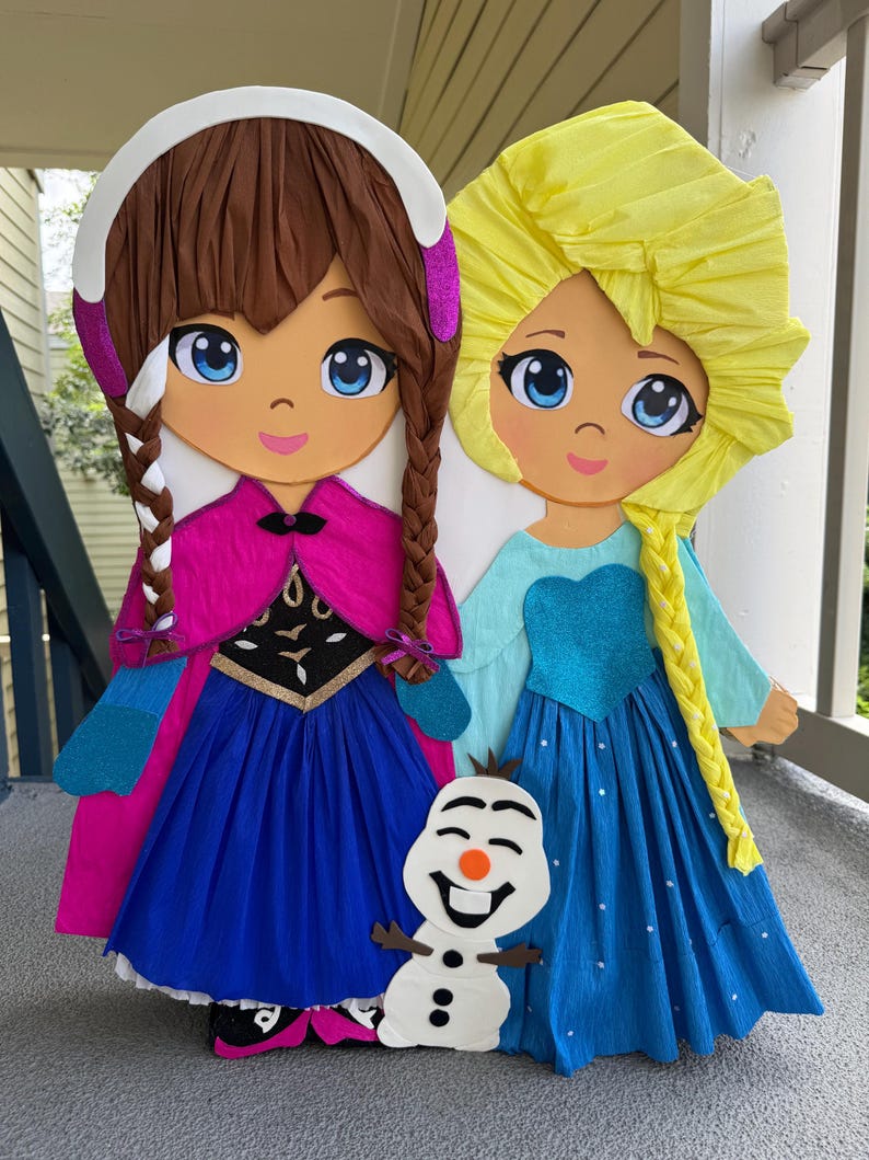 Puede incluir: Dos grandes mu&ntilde;ecas de papel de personajes animados, una con cabello casta&ntilde;o y una capa rosa, la otra con cabello amarillo y un vestido azul. Una peque&ntilde;a figura de mu&ntilde;eco de nieve est&aacute; frente a ellas. Las mu&ntilde;ecas est&aacute;n sobre una superficie gris.