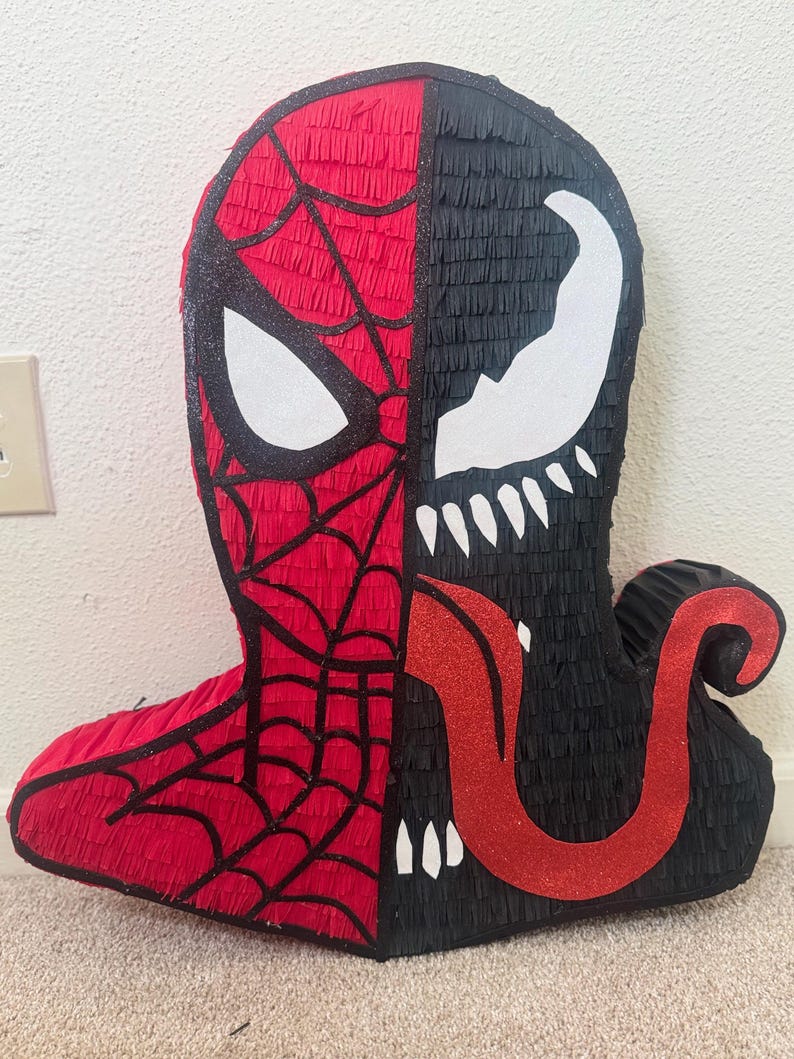 May include: A handmade pi&ntilde;ata featuring a split design of Spider-Man and Venom. The Spider-Man side is red with black webbing and a white eye. The Venom side is black with a white mouth and teeth, and a red glitter tongue.