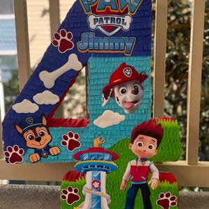 May include: A number four shaped piñata featuring the Paw Patrol logo, the name Jimmy, and character graphics including Marshall, Chase, Ryder, and the Lookout tower. The design incorporates blue, red, green, and white colors with paw print accents.