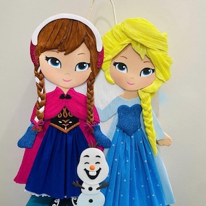 Puede incluir: Un adorno de pared decorativo con dos personajes de dibujos animados, uno con trenzas marrones y un vestido rosa y azul, el otro con una trenza amarilla y un vestido azul. Tambi&eacute;n se incluye una figura de mu&ntilde;eco de nieve sonriente.
