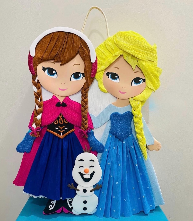 Puede incluir: Un adorno de pared decorativo con dos personajes de dibujos animados, uno con trenzas marrones y un vestido rosa y azul, el otro con una trenza amarilla y un vestido azul. Tambi&eacute;n se incluye una figura de mu&ntilde;eco de nieve sonriente.