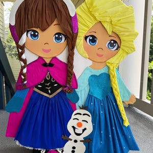 Puede incluir: Dos grandes y coloridas pi&ntilde;atas de personajes animados, una con cabello casta&ntilde;o y una capa rosa, la otra con cabello rubio y un vestido azul. Una peque&ntilde;a pi&ntilde;ata de mu&ntilde;eco de nieve est&aacute; colocada frente a ellas. Las pi&ntilde;atas est&aacute;n hechas de papel crep&eacute;.
