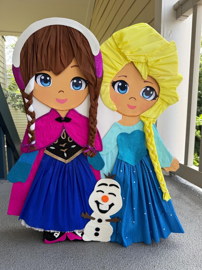 Puede incluir: Dos grandes y coloridas pi&ntilde;atas de personajes animados, una con cabello casta&ntilde;o y una capa rosa, la otra con cabello rubio y un vestido azul. Una peque&ntilde;a pi&ntilde;ata de mu&ntilde;eco de nieve est&aacute; colocada frente a ellas. Las pi&ntilde;atas est&aacute;n hechas de papel crep&eacute;.