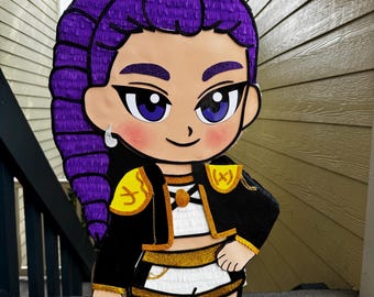 May include: A piñata shaped like a cartoon character with vibrant purple hair, large purple eyes, and a silver earring. The character wears a black and white outfit with gold glitter accents, including a cropped jacket, shorts, and tall black boots.
