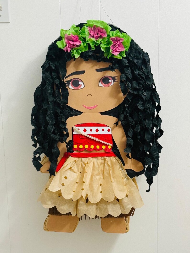 May include: A handmade pi&ntilde;ata in the shape of a cartoon character. The character has black paper hair, a floral crown, and a tan dress. The pi&ntilde;ata is designed with a red top and a tan skirt. The pi&ntilde;ata is designed to look like a cartoon character.