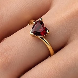 Puede incluir: Un anillo de oro con una piedra preciosa de color rojo oscuro en forma de corazón, montada en un engaste de garras. La banda es lisa y pulida, con la piedra en forma de corazón posicionada centralmente en el dedo. El diseño es sencillo, destacando el contraste entre el metal dorado y el color rojo intenso de la piedra tallada.