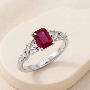 May include: A silver ring featuring a rectangular, deep red gemstone as the centerpiece. The ring is adorned with smaller, clear gemstones and decorative leaf-like accents on the band, creating an elegant and sophisticated design.