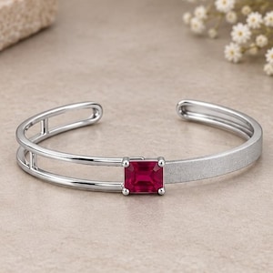 Peut inclure: Un bracelet manchette argenté orné d'une pierre précieuse rectangulaire rouge foncé au centre. Le bracelet présente un design à double rail d'un côté et une finition mate texturée de l'autre, créant un contraste entre les surfaces lisses et rugueuses.