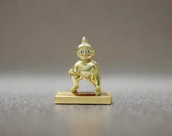 Può includere: Una statuetta dorata di una divinità in posizione accovacciata, montata su una base rettangolare. La figura presenta lineamenti dettagliati ed è probabilmente un manufatto religioso o culturale. Lo sfondo è grigio neutro.