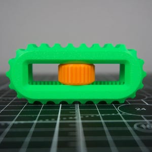 May include: A bright green, rectangular 3D-printed object with a ridged edge and a central orange cylinder. The object has a geometric design and appears to be a tool or component.