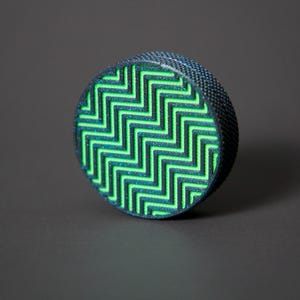 May include: A round, textured object with a black exterior and a neon green zigzag pattern on the top. The object is set against a dark gray background, creating a striking contrast. The design is geometric and eye-catching.
