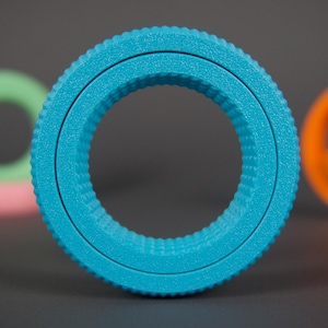 May include: A close-up of a vibrant turquoise ring with a textured, ridged outer edge. Other rings in shades of green, pink, and orange are blurred in the background, creating a colorful, abstract composition.