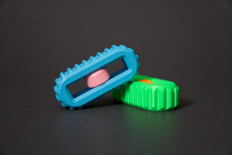 May include: Two colorful, geometric toys. One is light blue with a pink center and a ridged edge. The other is green with an orange accent. The toys are on a dark gray surface.