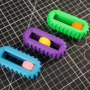 May include: Three colorful 3D-printed objects. Each has a rectangular frame with a toothed edge and a cylindrical center. The objects are teal, purple, and green, with orange, yellow, and pink centers, respectively. They rest on a black cutting mat.