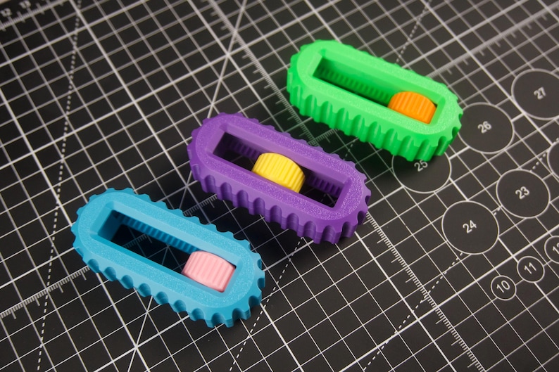 May include: Three colorful 3D-printed objects. Each has a rectangular frame with a toothed edge and a cylindrical center. The objects are teal, purple, and green, with orange, yellow, and pink centers, respectively. They rest on a black cutting mat.